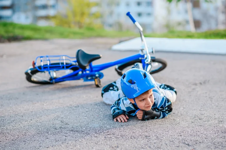 Šioje nuotraukoje vaizduojamas mažas vaikas, kuris nukrito nuo dviračio. Jis guli ant pilvo ant asfalto, apsirengęs apsaugine apranga, įskaitant šalmą ir apsaugas ant rankų bei kojų. Mėlynas dviratis guli ant šono už jo. Fone – pastatas ir žolė.