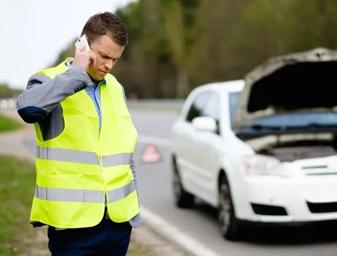 Sugedęs baltas automobilis pakelėje: vairuotojas su atšvaitine liemene skambina techninei pagalbai.
